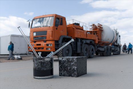 Un bécher de bitume et une éprouvette d'enrobé sur le chantier, des engins routiers derrière